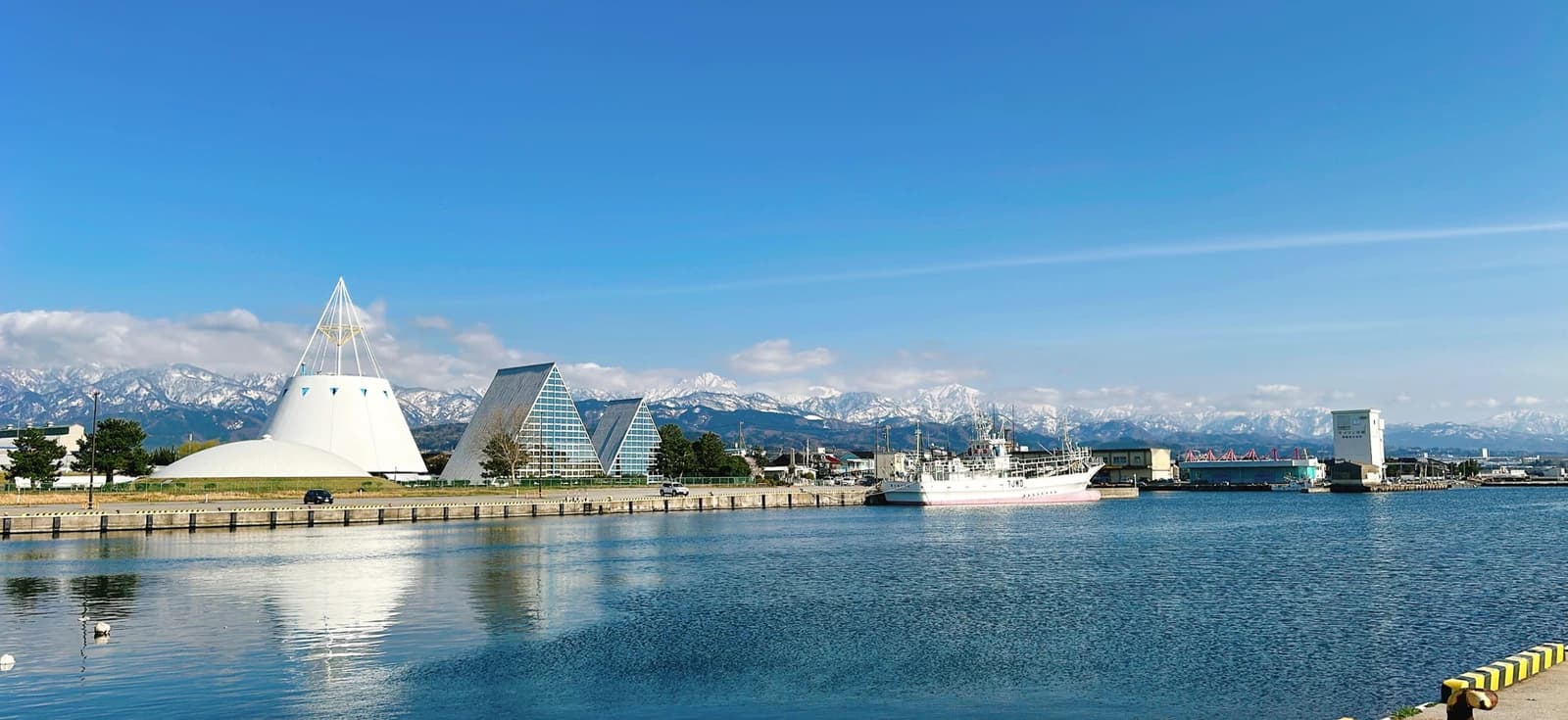 晴れた日の魚津港。港の岸壁と停泊する漁船の向こうに、とんがり屋根の建物と雪をかぶった立山連峰が見える