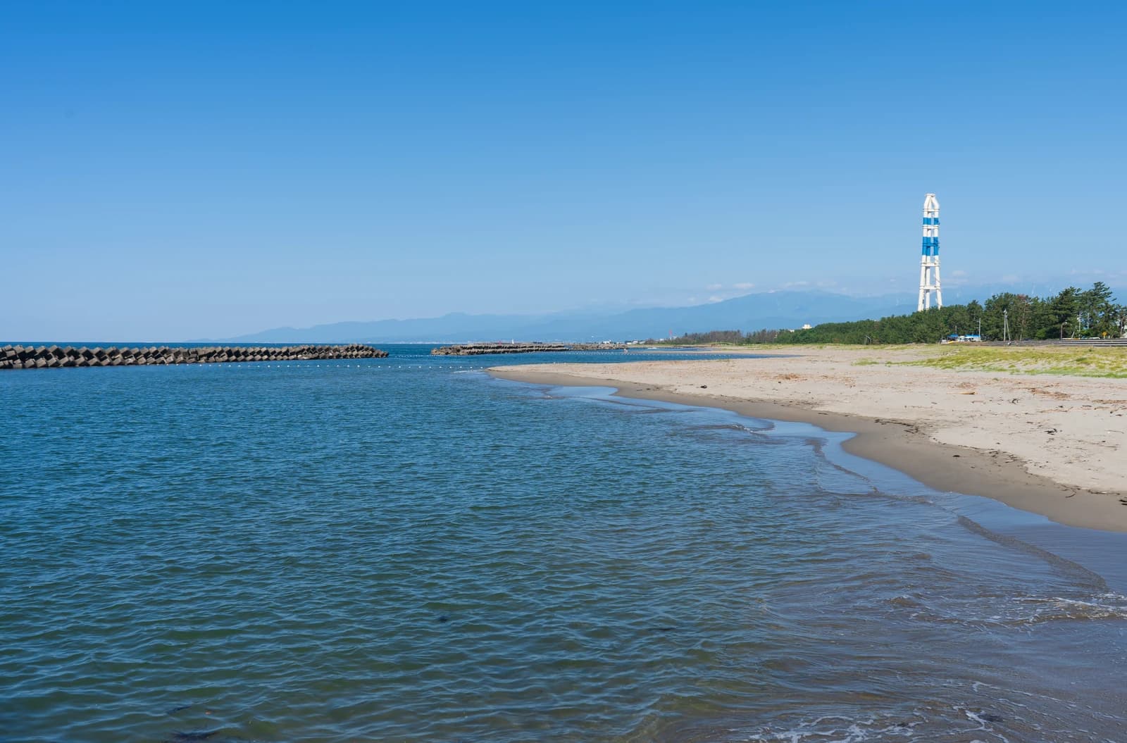 晴れた日の八重津浜。遠浅の海と砂浜が続き、奥に白と青の鉄塔と松林、立山連峰が見える