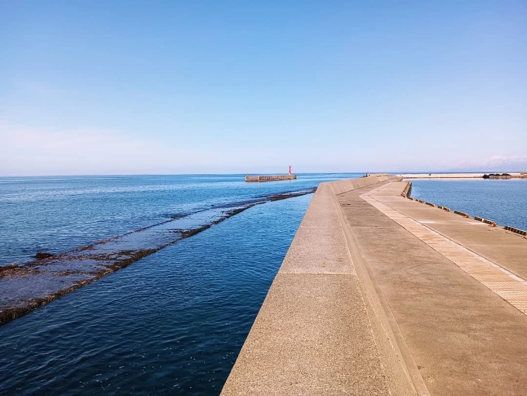 晴れた日の四方漁港。海に沿って延びる防波堤と、その先の赤い灯台